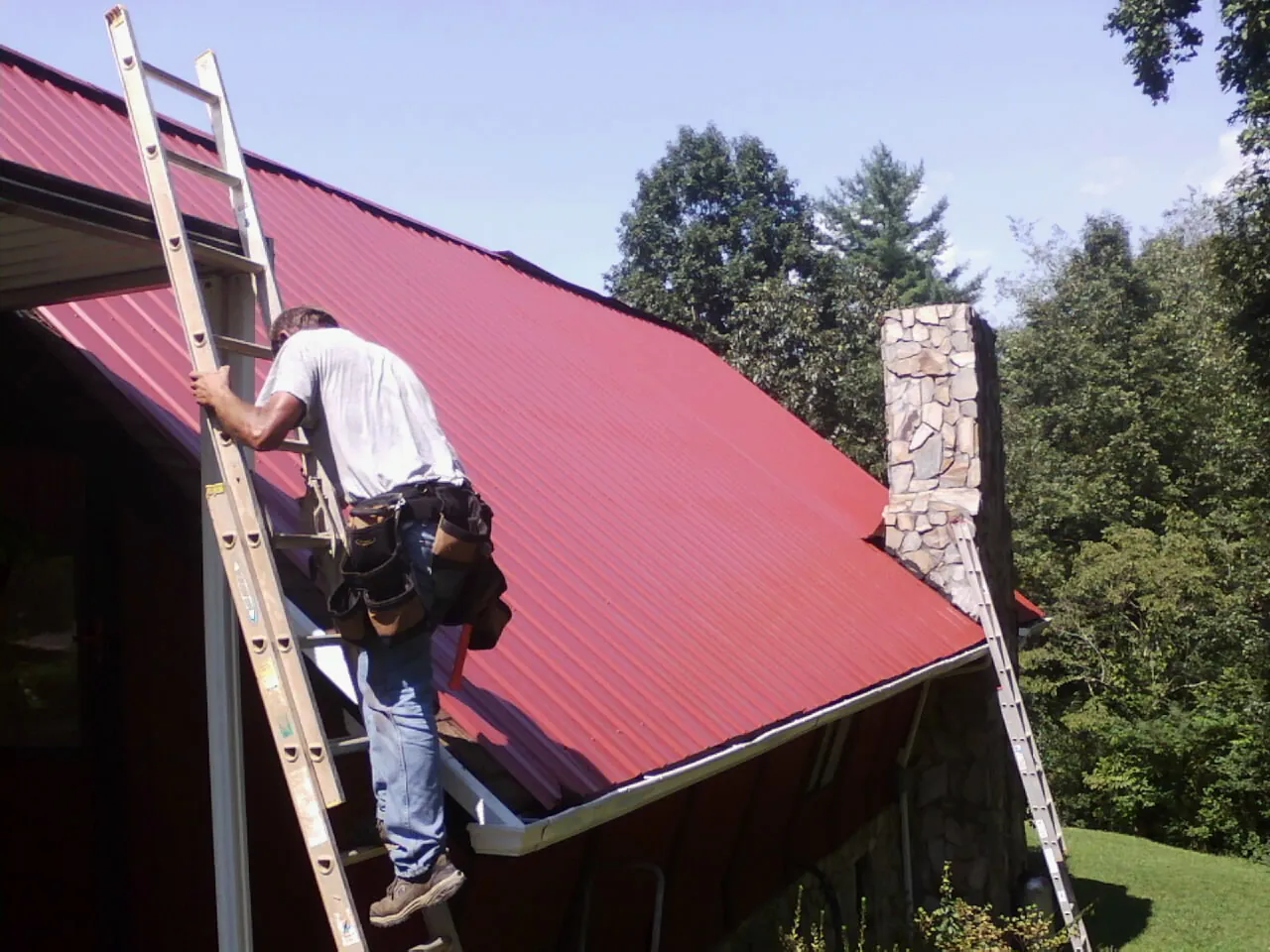 Roofer on a ladder beside a red metal roof and stone chimney — Soffit & Fascia Repair by Roofing Brothers in Skaneateles Falls
